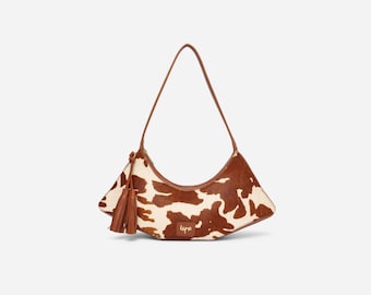 May include: A crescent-shaped shoulder bag featuring a brown and white cowhide pattern. The bag includes a thin brown leather strap, a matching brown leather tassel attached to the side, and a small gold-toned logo plate on the front surface.