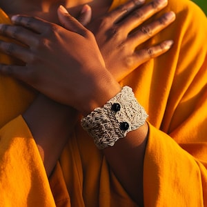 Puede incluir: Una pulsera tejida con dos cuentas negras se lleva en la muñeca. La pulsera está hecha de un material claro y texturizado. El fondo es una tela de color amarillo-naranja brillante.