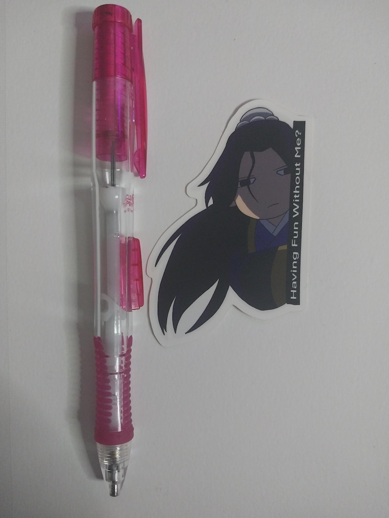 May include: A clear and pink mechanical pencil with a pink grip and accents, next to a sticker featuring a cartoon character with long black hair and the text "Having Fun Without Me?"