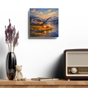 Puede incluir: Un reloj de pared cuadrado con una pintura impresionista de una furgoneta camper blanca aparcada junto a un lago al atardecer. Una hoguera brillante resplandece en la orilla, reflejándose en el agua. El reloj tiene manecillas negras.