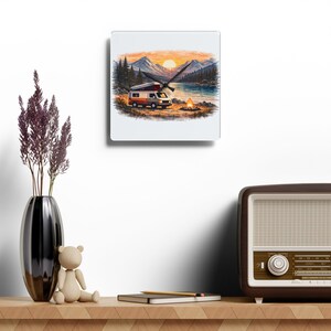 Puede incluir: Un reloj de pared cuadrado con una ilustración escénica de una furgoneta camper junto a un lago y montañas al atardecer. La esfera del reloj muestra una fogata. Las manecillas del reloj son negras.