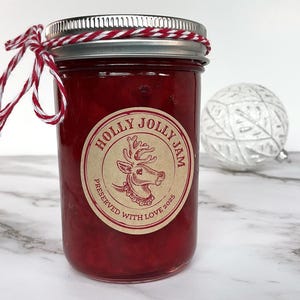 Puede incluir: Un frasco de vidrio lleno de mermelada roja, con una tapa de metal plateado y un lazo de cuerda roja y blanca. La etiqueta circular presenta una ilustración de un reno y el texto HOLLY JOLLY JAM PRESERVED WITH LOVE 2025 en tinta roja.