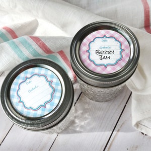 May include: Two glass jars with metal lids. The lids have round, blue and white gingham patterned labels with a white frame and the words "Date:" and "Contents:". One jar has a pink and white gingham label with the words "Berry Jam".