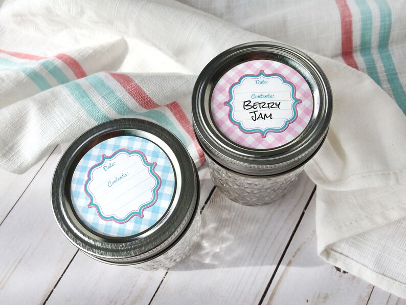 May include: Two glass jars with metal lids. The lids have round, blue and white gingham patterned labels with a white frame and the words "Date:" and "Contents:". One jar has a pink and white gingham label with the words "Berry Jam".