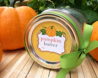 May include: A glass jar with a green ribbon tied around the lid. The jar has a yellow and white polka dot label that says "pumpkin butter" with a small pumpkin image. The jar is sitting on a brown woven mat with two orange pumpkins in the background.