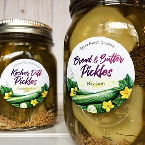 May include: Two glass jars of homemade pickles. One jar is labeled "Kosher Dill Pickles" and the other "Bread & Butter Pickles." Both jars have decorative labels with cucumber and flower illustrations. The pickles are canned in 2020.