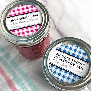 May include: Two glass jars of homemade jam with metal lids. One jar is labeled "Raspberry Jam From Alison's Kitchen" with a pink gingham pattern. The other jar is labeled "Maine's Finest Blueberry Jam From Tim's Backyard" with a blue gingham pattern.