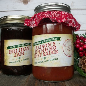 May include: Two glass jars with red and green holiday themed labels. The jar on the left is filled with a dark brown liquid and labeled "Happy Holidays! Holiday Jam* From: Fred & Cheryl". The jar on the right is filled with a red liquid and labeled "Merry Christmas Alison's Ho Ho Ho Hot Sauce* Eat, Drink, & Be Merry! Made with Love Date 2019".