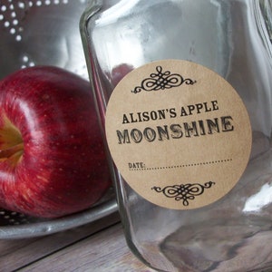 May include: A clear glass bottle with a brown paper label that says "Alison's Apple Moonshine" and has a space for a date. The label has a decorative design at the top and bottom. Two red apples are in the background.