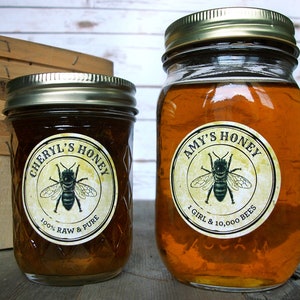 May include: Two glass jars of honey with gold lids. The jars have round labels with a bee illustration and text that reads "Cheryl's Honey 100% Raw & Pure" and "Amy's Honey 1 Girl & 10,000 Bees".