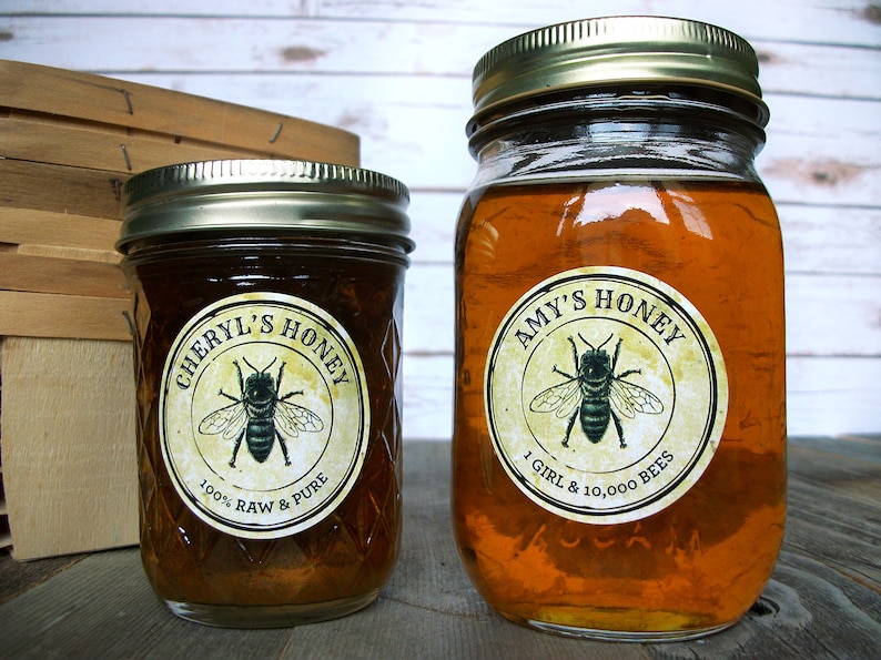 May include: Two glass jars of honey with gold lids. The jars have round labels with a bee illustration and text that reads "Cheryl's Honey 100% Raw & Pure" and "Amy's Honey 1 Girl & 10,000 Bees".