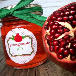 May include: A glass jar filled with red pomegranate jelly, topped with a green ribbon and a white label with red polka dots. The label reads "pomegranate jelly" and has a small illustration of a pomegranate. A half of a pomegranate is visible in the background.