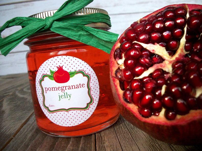 May include: A glass jar filled with red pomegranate jelly, topped with a green ribbon and a white label with red polka dots. The label reads "pomegranate jelly" and has a small illustration of a pomegranate. A half of a pomegranate is visible in the background.