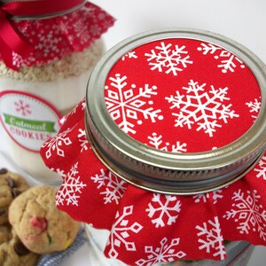 Close-up of a glass jar with a metal lid, covered with a red fabric featuring white snowflakes. The jar is filled with a light-colored mixture, and a label reads "Oatmeal Cookies". A few cookies are visible in the foreground.