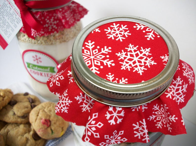 Close-up of a glass jar with a metal lid, covered with a red fabric featuring white snowflakes. The jar is filled with a light-colored mixture, and a label reads "Oatmeal Cookies". A few cookies are visible in the foreground.