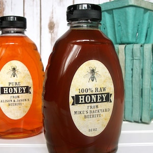 Vintage looking oval honey labels on the front of honey bottles filled with honey. Label art has a realistic honey bee up top with printed text for 100% Raw honey. In the background are green berry baskets.
