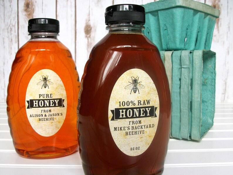 Vintage looking oval honey labels on the front of honey bottles filled with honey. Label art has a realistic honey bee up top with printed text for 100% Raw honey. In the background are green berry baskets.