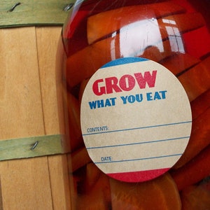 May include: A glass jar filled with orange carrot slices in a red liquid. A round label with blue and red trim reads "GROW WHAT YOU EAT". The label has blank spaces for "CONTENTS" and "DATE".