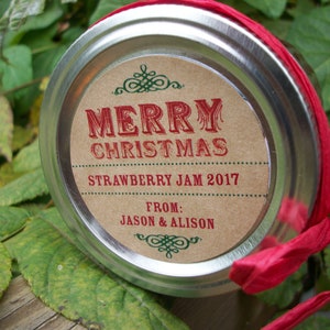 Puede incluir: Una tapa de tarro redonda de metal plateado con una etiqueta circular de color canela con texto en rojo y verde que dice MERRY CHRISTMAS STRAWBERRY JAM 2017 FROM: JASON & ALISON. Una cinta de rafia roja brillante rodea la tapa.