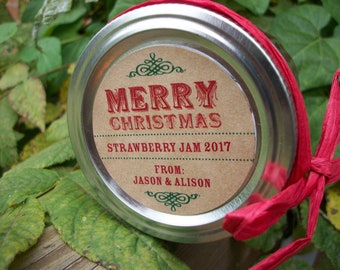 May include: A round silver metal jar lid featuring a tan circular label with red and green text that reads MERRY CHRISTMAS STRAWBERRY JAM 2017 FROM: JASON & ALISON. A bright red raffia ribbon is tied around the side of the metal lid.