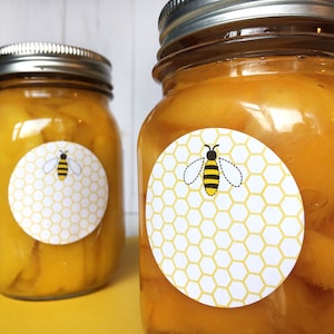May include: Two glass jars filled with a yellow substance, likely honey or jam, with white round labels featuring a yellow and black bee on a honeycomb pattern.