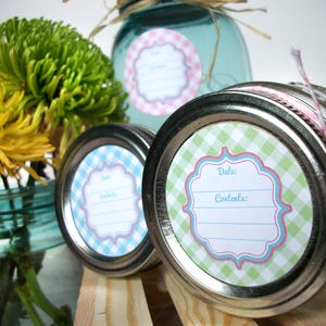 May include: Three glass jars with lids, each with a different coloured gingham patterned label. The labels have a space for "Date" and "Contents". The jars are on a wooden surface with flowers in the background.