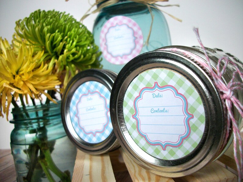 May include: Three glass jars with lids, each with a different coloured gingham patterned label. The labels have a space for "Date" and "Contents". The jars are on a wooden surface with flowers in the background.