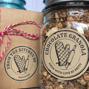 May include: A brown paper gift box tied with red and white twine. The box has a round sticker that says "From the Kitchen of Alison Kay" with a drawing of a kitchen mitt and rolling pin. A glass jar filled with chocolate granola has a round sticker that says "Chocolate Granola Made with Love by Patty" with a drawing of a kitchen mitt and rolling pin.