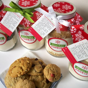 Several mason jars filled with cookie ingredients, labeled "Oatmeal Cookies", "Candy Chip Cookies", "Peanut Butter Cookies", and "Chocolate Chip Cookies". Each jar has a red and white snowflake lid, a recipe tag, and a ribbon. A plate of cookies sits in front.