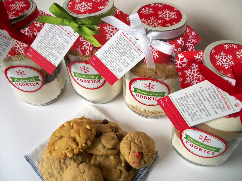 Several mason jars filled with cookie ingredients, labeled "Oatmeal Cookies", "Candy Chip Cookies", "Peanut Butter Cookies", and "Chocolate Chip Cookies". Each jar has a red and white snowflake lid, a recipe tag, and a ribbon. A plate of cookies sits in front.