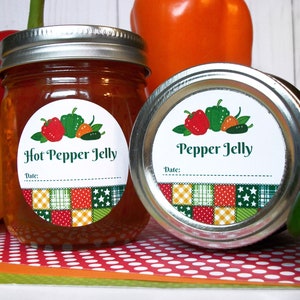 May include: Two glass jars with silver lids and round labels. The labels are white with green text and a patchwork design. The labels say "Hot Pepper Jelly" and "Pepper Jelly". The jars are surrounded by red, orange, and green peppers.