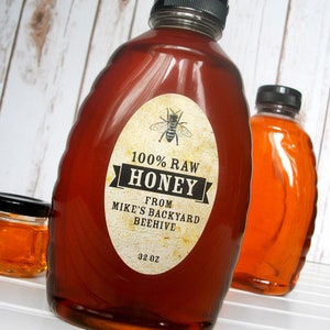 Vintage looking oval honey label on the front of honey bottle filled with honey. Label art has a realistic honey bee up top with printed text for 100% Raw honey. In the background are jars filled with honey.