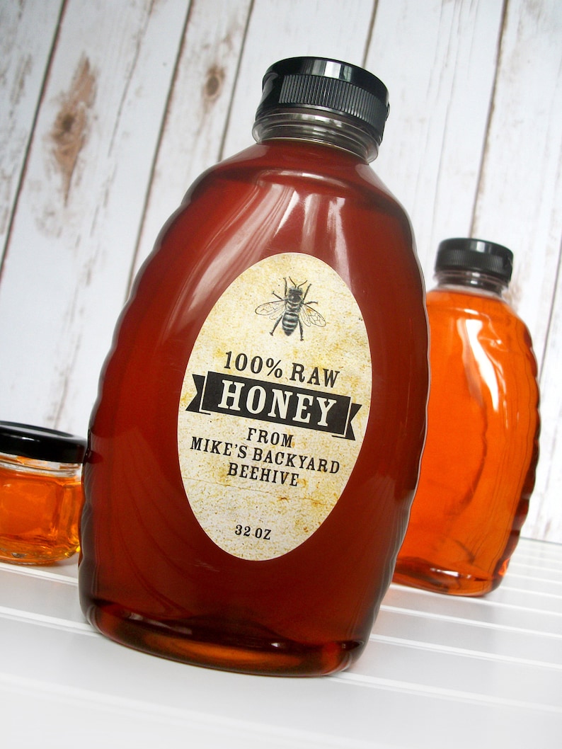 Vintage looking oval honey label on the front of honey bottle filled with honey. Label art has a realistic honey bee up top with printed text for 100% Raw honey. In the background are jars filled with honey.