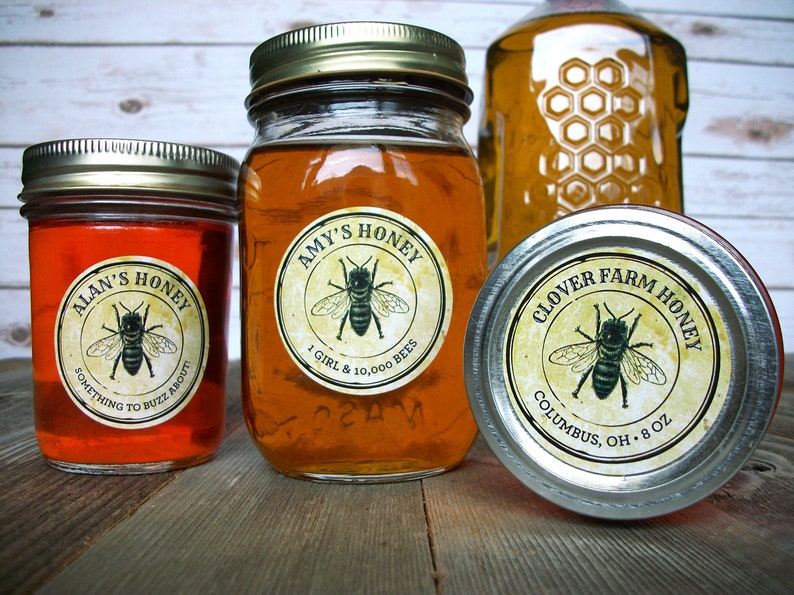 May include: Three glass jars of honey with labels. The labels are round and have a vintage design with a bee illustration. The labels read "Alan's Honey", "Amy's Honey", and "Clover Farm Honey".