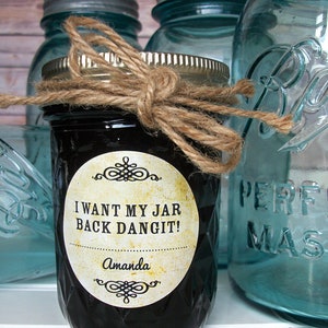 May include: A glass jar with a black liquid and a label that reads "I want my jar back dangit!" with the name Amanda below. The jar is tied with twine and is sitting on a white surface with other glass jars in the background.