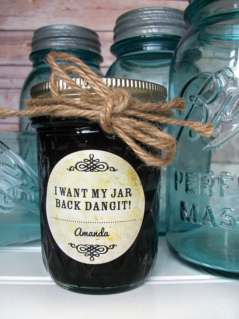 May include: A glass jar with a black liquid and a label that reads "I want my jar back dangit!" with the name Amanda below. The jar is tied with twine and is sitting on a white surface with other glass jars in the background.