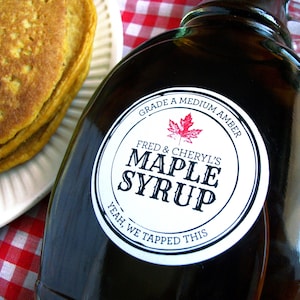 May include: A glass bottle of maple syrup with a white label that reads "Grade A Medium Amber Fred & Cheryl's Maple Syrup Yeah, We Tapped This". The label has a red maple leaf and a black circle around the text.