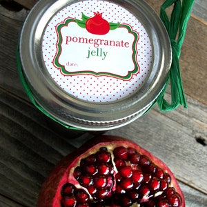May include: A silver jar with a white label that reads "pomegranate jelly" with a red and green illustration of a pomegranate. A half of a pomegranate with red seeds is in the foreground.