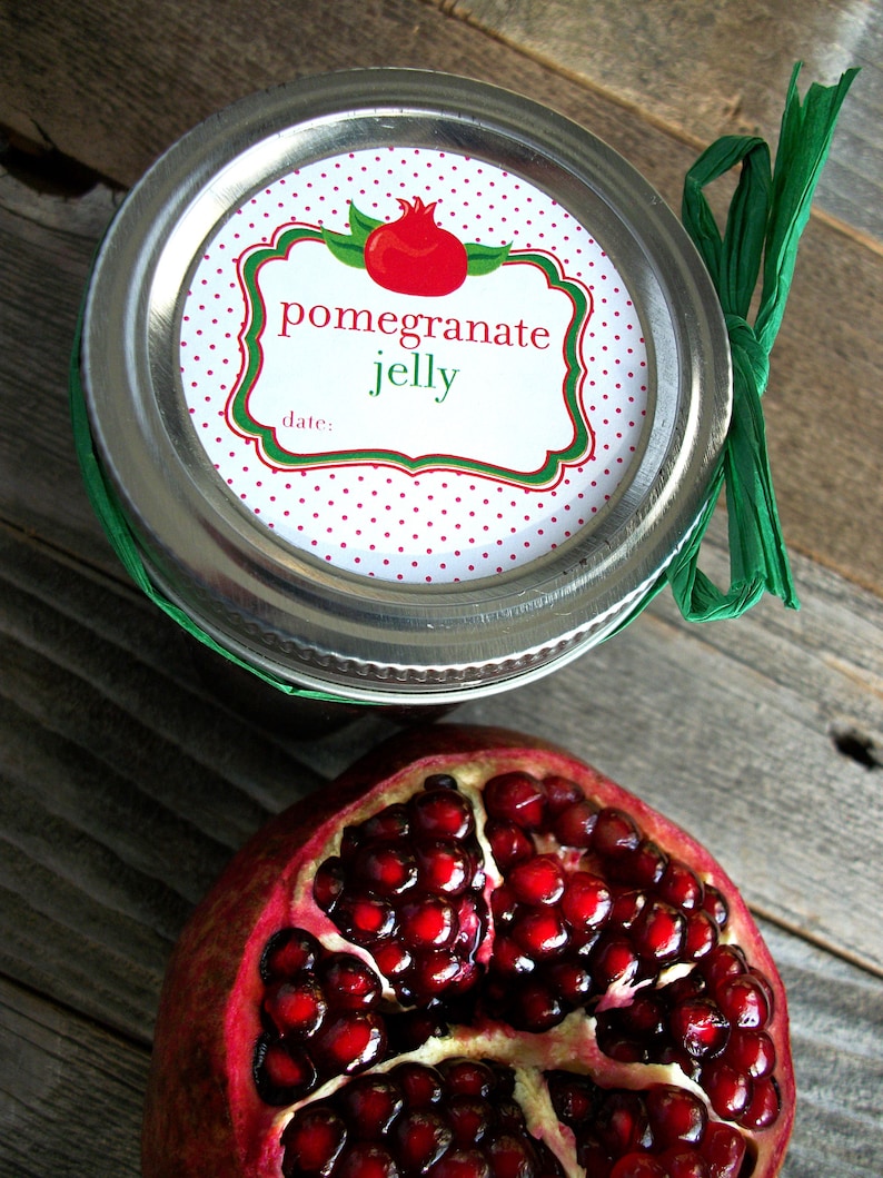 May include: A silver jar with a white label that reads "pomegranate jelly" with a red and green illustration of a pomegranate. A half of a pomegranate with red seeds is in the foreground.