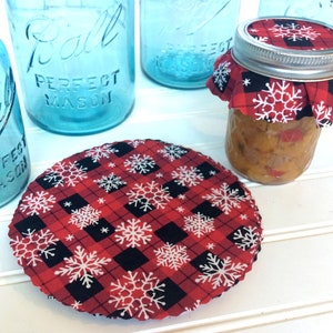 Puede incluir: Tela de cuadros rojos y negros con copos de nieve blancos impresos en ella. La tela tiene forma circular y se utiliza como tapa para un frasco de vidrio.