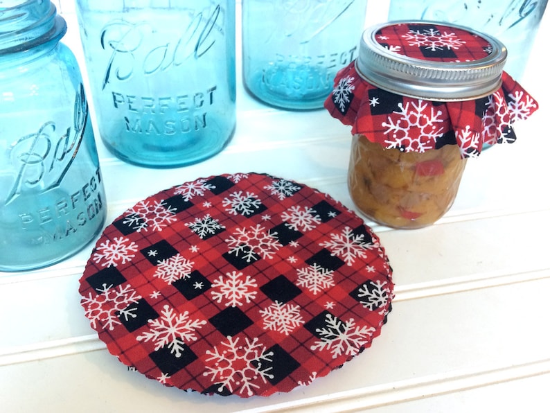 Puede incluir: Tela de cuadros rojos y negros con copos de nieve blancos impresos en ella. La tela tiene forma circular y se utiliza como tapa para un frasco de vidrio.