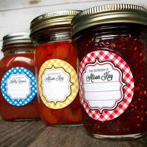 May include: Three glass jars with red, yellow, and red and white gingham patterned labels. The labels have a white background with black text that reads "From the Kitchen of" followed by a name. The jars contain red sauce, peppers, and jam.