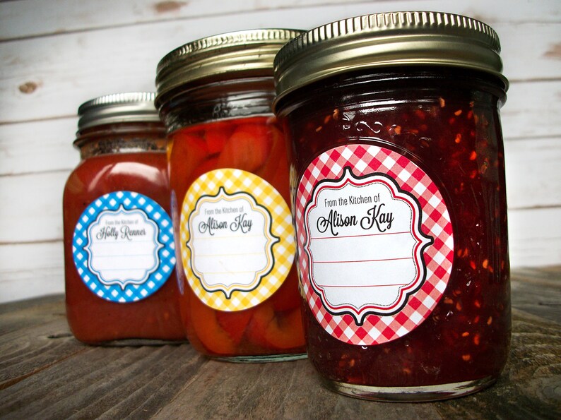 May include: Three glass jars with red, yellow, and red and white gingham patterned labels. The labels have a white background with black text that reads "From the Kitchen of" followed by a name. The jars contain red sauce, peppers, and jam.