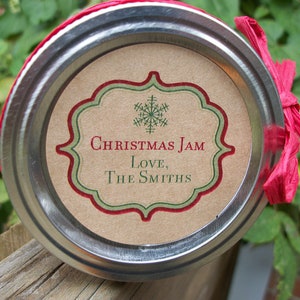 Puede incluir: Una tapa de frasco redonda de metal con una etiqueta de papel color canela, un icono de copo de nieve verde, un borde decorativo rojo y verde, y el texto "Christmas Jam Love, The Smiths". Una cinta de rafia rosa brillante está atada al lado.