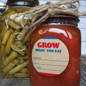 May include: Two glass jars with lids, one filled with green beans and the other with red salsa. The salsa jar has a brown paper label with the text "GROW WHAT YOU EAT" in red and blue letters. The label also has blank spaces for "CONTENTS" and "DATE".