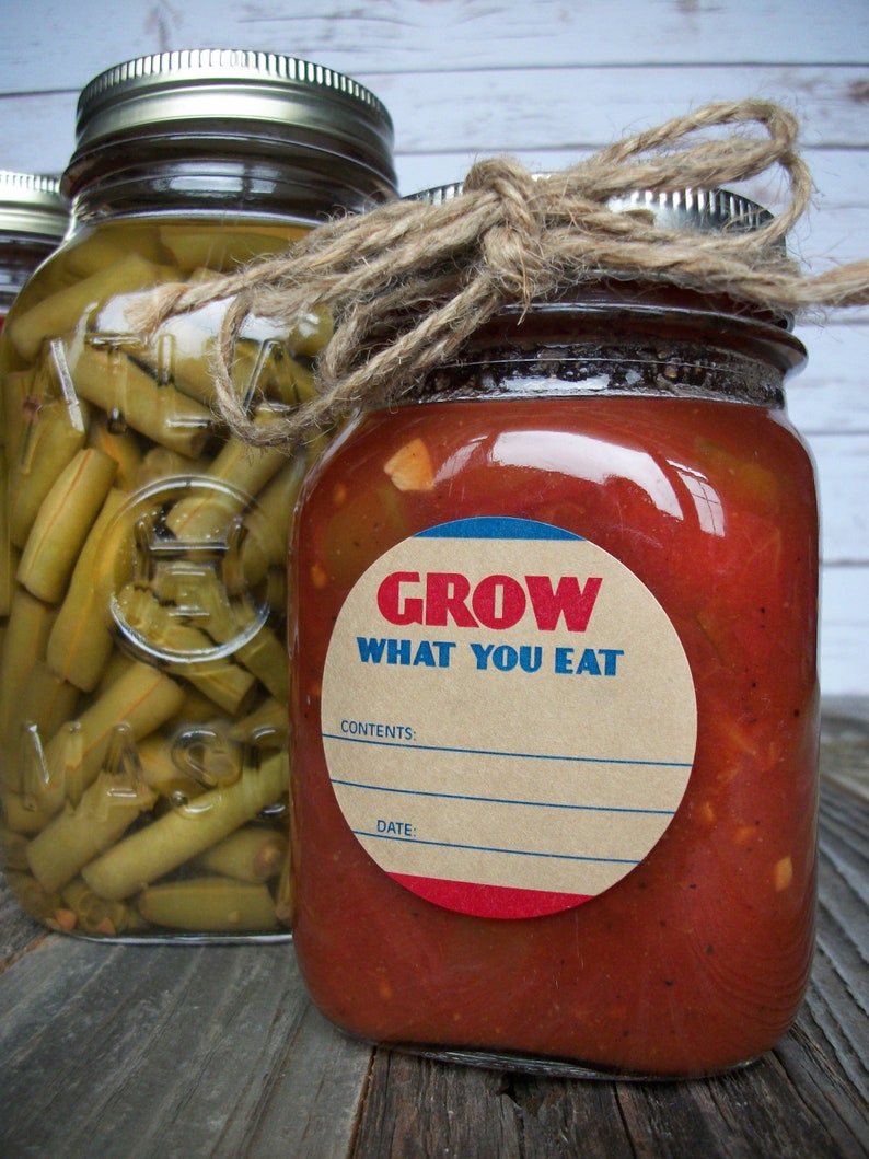 May include: Two glass jars with lids, one filled with green beans and the other with red salsa. The salsa jar has a brown paper label with the text "GROW WHAT YOU EAT" in red and blue letters. The label also has blank spaces for "CONTENTS" and "DATE".