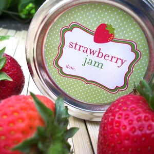 May include: A green and white polka dot jar lid with a white label that says "strawberry jam" in red and green lettering. The label has a green and red border and a space for a date. There are three red strawberries in the image.