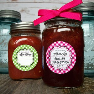May include: Two glass jars with lids, one filled with red sauce and the other with pink jam. Both jars have round, white and green gingham and white and pink gingham labels with black text. The label on the jar of sauce reads "From the Kitchen of Alan & Peggy". The label on the jar of jam reads "From the Kitchen of Alison Kay Berry Preserves 2018".