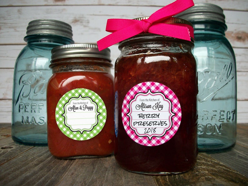 May include: Two glass jars with lids, one filled with red sauce and the other with pink jam. Both jars have round, white and green gingham and white and pink gingham labels with black text. The label on the jar of sauce reads "From the Kitchen of Alan & Peggy". The label on the jar of jam reads "From the Kitchen of Alison Kay Berry Preserves 2018".