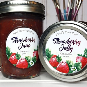 May include: Two glass jars of homemade strawberry jam and jelly. The jam jar has a label that reads "Strawberry Jam, Summer 2020, From Alison's Kitchen." The jelly jar lid reads "Strawberry Jelly, Canned by Janet, It's Jelly Time, Y'all!"